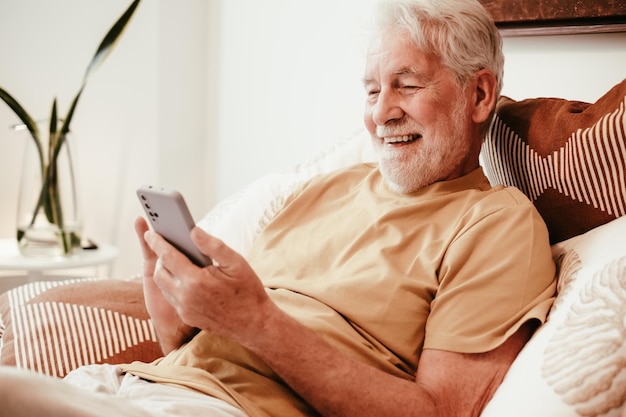 Retrato de un hombre mayor feliz, relajado y guapo en la cama sosteniendo un teléfono inteligente, un abuelo jubilado usando aplicaciones telefónicas
