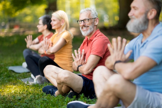 Foto retrato de un hombre mayor guapo que asiste a clases de yoga en grupo al aire libre, un caballero maduro feliz practicando meditación con amigos en el parque, sentado en posición de loto y sonriendo a la cámara