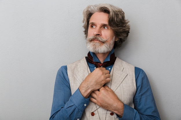 Retrato de un hombre mayor guapo serio barbudo con cabello gris aislado sobre fondo gris de la pared mirando a un lado.