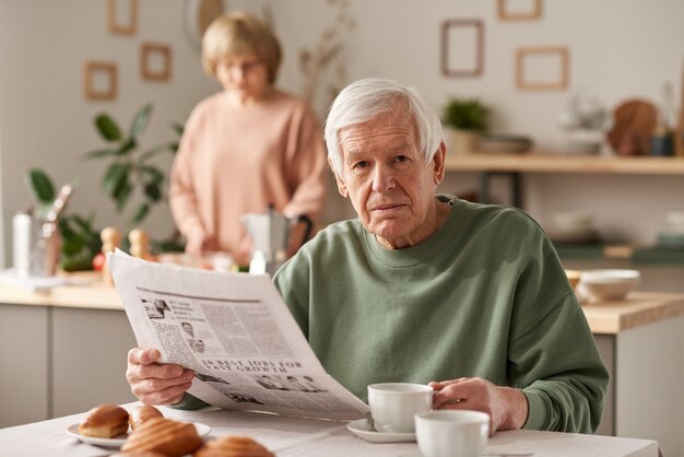 Retrato de hombre mayor mirando a la cámara mientras lee el periódico durante el desayuno en la cocina