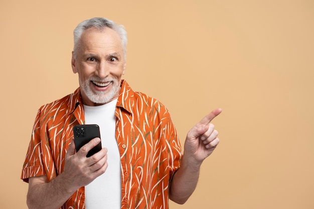Foto retrato de un hombre mayor sonriente y emocionado de pelo gris sosteniendo un teléfono móvil usando un teléfono inteligente apuntando con el dedo al espacio de copia aislado sobre fondo beige un pensionista atractivo mirando la cámara