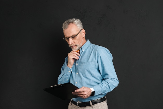 Retrato de hombre de mediana edad de 60 años con canas y barba trabajando con documentos mientras sostiene el portapapeles, aislado sobre la pared negra