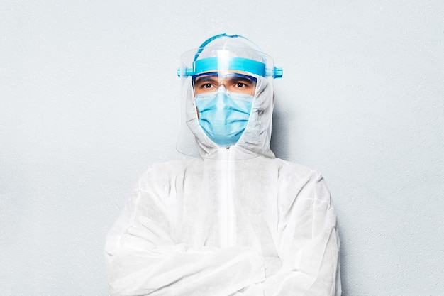 Retrato de hombre médico con los brazos cruzados, vestido con traje PPE contra coronavirus y covid-19, en la pared de la pared con textura blanca.