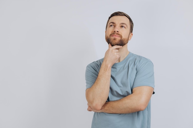 Retrato de un hombre moderno de mediana edad con barba en una camiseta azul que muestra emoción pensando en algo sobre un fondo blanco