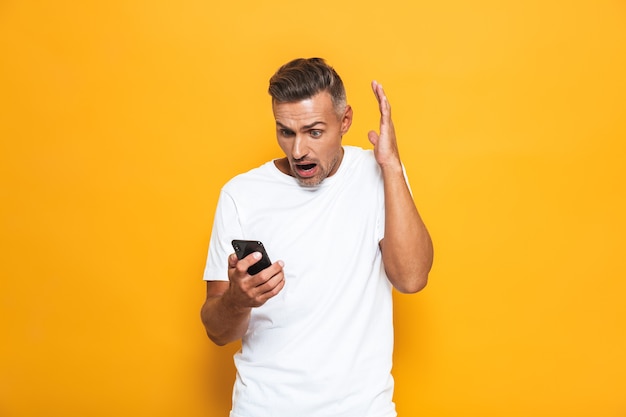 Retrato de hombre molesto de 30 años en camiseta blanca gritando y sosteniendo el teléfono inteligente mientras está de pie aislado en amarillo