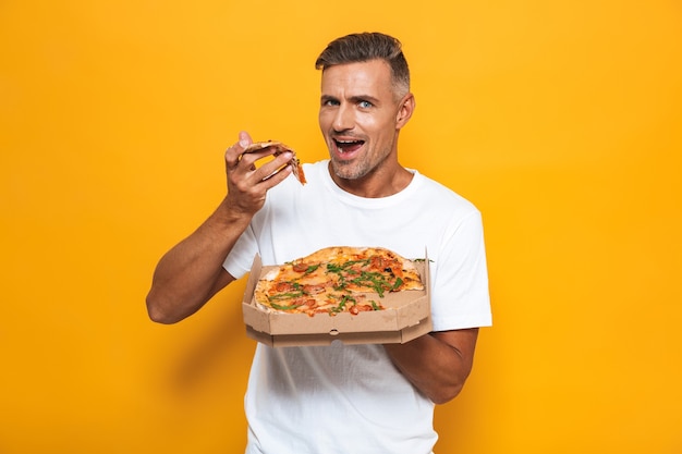 Retrato de hombre morena de 30 años en camiseta blanca sosteniendo y comiendo pizza mientras está de pie aislado en amarillo