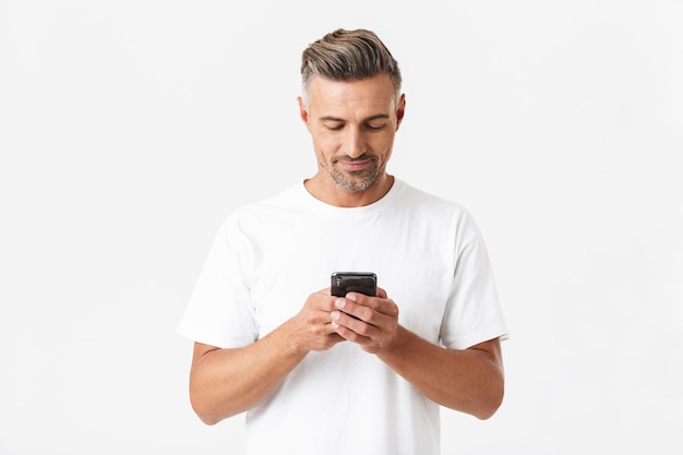 Retrato de hombre morena de 30 años con camiseta casual sosteniendo y usando smartphone aislado en blanco