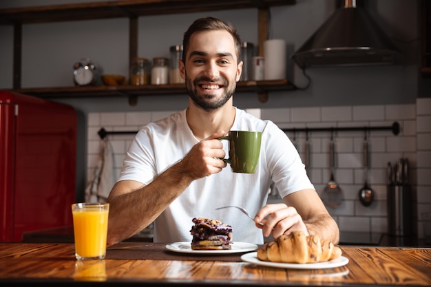 Retrato de hombre morena de 30 años comiendo croissants y bebiendo jugo mientras desayuna en la elegante cocina de casa