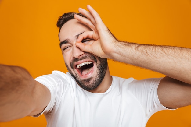 Foto retrato de hombre morena en camiseta sonriendo mientras toma una foto selfie, aislado en amarillo