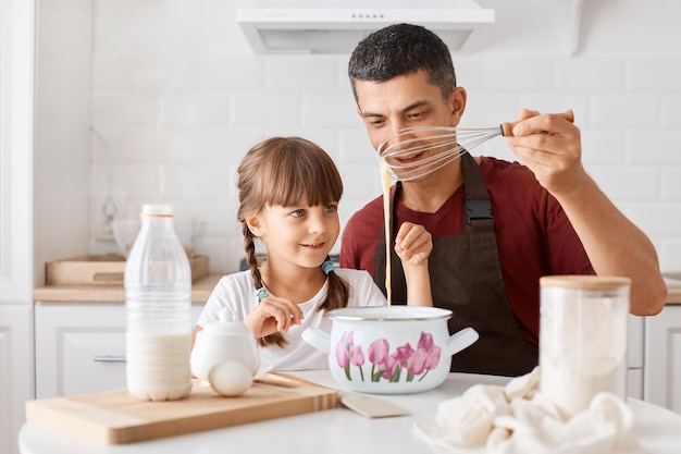 Retrato de un hombre moreno con delantal sentado en la mesa con su hija y cocinando juntos en la cocina mezclando masa para panqueques horneando postre sabroso