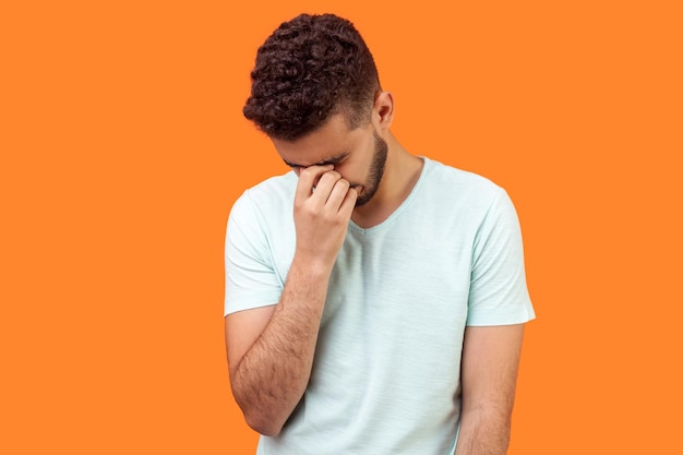 Retrato de un hombre moreno solitario deprimido con barba en una camiseta blanca casual sosteniendo la cabeza hacia abajo, escondiendo su rostro en la mano y llorando, sintiéndose desesperado. tiro de estudio interior aislado sobre fondo naranja