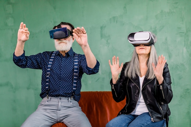 Retrato de hombre y mujer de edad avanzada con estilo, vistiendo ropa de moda, sentado en una silla roja con gafas de realidad virtual y tocando una pantalla imaginaria en el aire