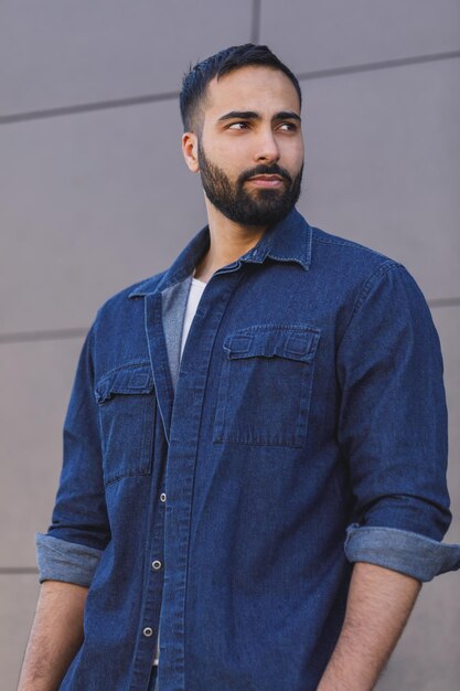 Retrato de un hombre multicultural confiado y barbudo con camisa de jeans parado cerca del edificio moderno