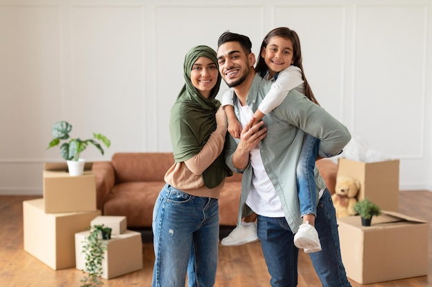 Retrato de hombre musulmán emocionado, mujer con pañuelo en la cabeza y niña posando en apartamento nuevo, de pie en la sala de estar vacía con cajas de cartón. Padre alegre con su hija en la espalda