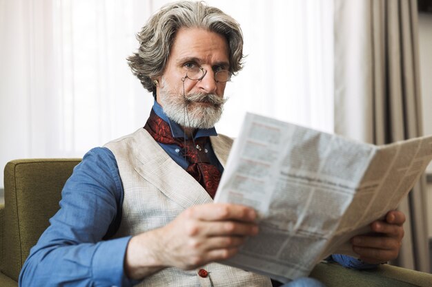 Retrato de hombre de negocios adulto barbudo vistiendo elegante traje leyendo el periódico mientras está sentado en un sillón en el apartamento del hotel