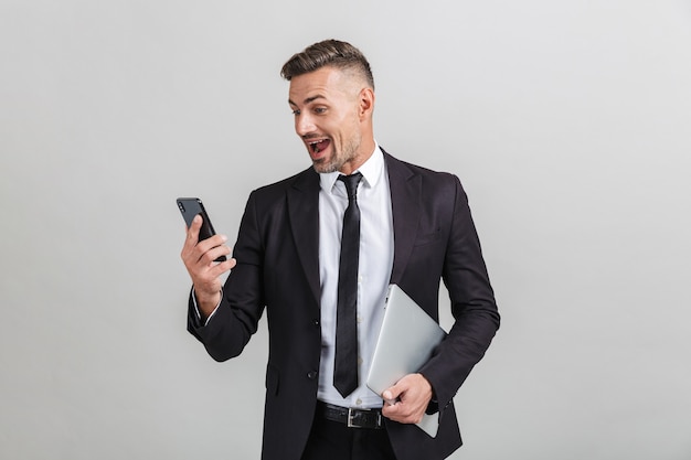 Retrato de hombre de negocios adulto emocionado en traje de oficina mirando el teléfono inteligente y sosteniendo la computadora portátil mientras está de pie aislado