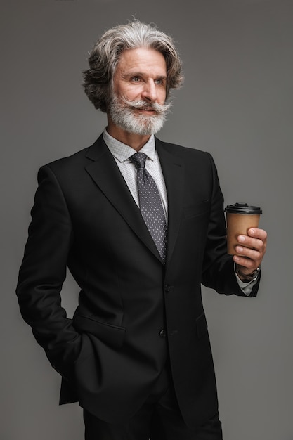 Retrato de hombre de negocios adulto vistiendo traje negro formal sonriendo y sosteniendo un vaso de papel aislado sobre pared gris