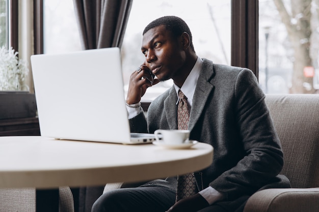 Retrato del hombre de negocios africano feliz que usa el teléfono mientras que trabaja en la computadora portátil en un restaurante.