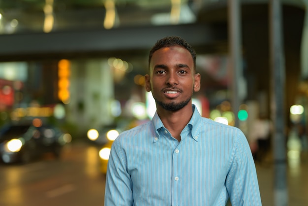 Retrato de hombre de negocios africano negro guapo al aire libre en la ciudad por la noche sonriendo tiro horizontal