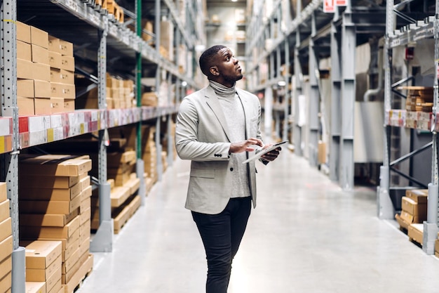 Retrato de un hombre de negocios afroamericano sonriente que ordena detalles en tabletas que verifican bienes y suministros en estantes con antecedentes de bienes en almacén. Logística y exportación comercial