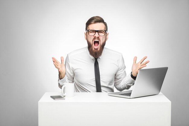 Retrato de hombre de negocios agresivo con camisa blanca y corbata negra está sentado en la oficina y tiene mal humor con los brazos levantados mirando a la cámara y gritando. tiro del estudio de interior, fondo gris aislado