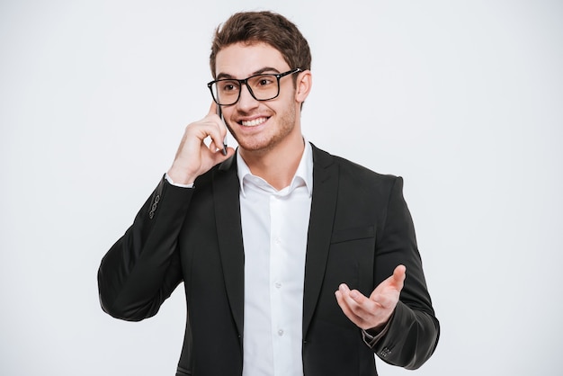 Retrato de un hombre de negocios alegre en anteojos hablando por teléfono aislado en una pared blanca