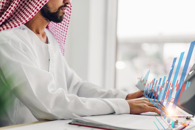 Retrato de un hombre de negocios árabe de éxito árabe que utiliza la tecnología de una computadora portátil que observa el progreso del gráfico de datos de inversión financiera empresarial y el aumento del crecimiento empresarial en la oficina
