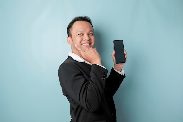 Un retrato de un hombre de negocios asiático feliz sonríe y sostiene su teléfono inteligente señalando el espacio de copia a su lado con traje negro aislado por un fondo azul