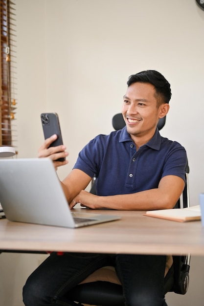 Retrato de un hombre de negocios asiático feliz usando su teléfono inteligente en su escritorio de oficina