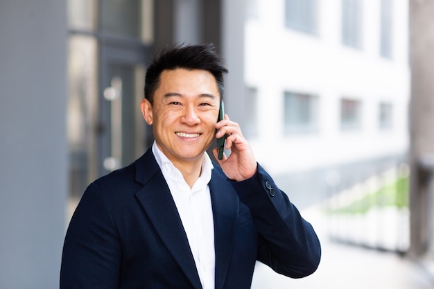 Retrato de hombre de negocios asiático hablando por teléfono alegre sonriente informes buenas noticias cerca del centro de la oficina exterior mirando a la cámara