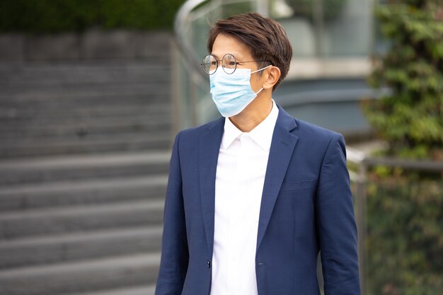 Retrato de hombre de negocios asiático con mascarilla protectora para prevenir el virus covid-19 de pie al aire libre en la ciudad.