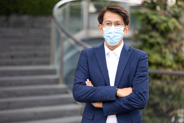 Retrato de hombre de negocios asiático con mascarilla protectora para prevenir el virus covid-19 de pie al aire libre en la ciudad.