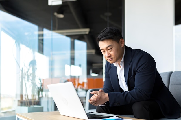 Retrato de un hombre de negocios asiático pensativo que trabaja en una computadora portátil en un escritorio de oficina moderno Hombre pensativo enfocado confiado en traje formal en el interior pensando en la inspiración para resolver un problema Inicio