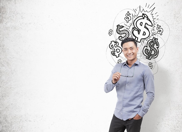Retrato de un hombre de negocios asiático sonriente que lleva una camisa azul y pantalones oscuros y gafas de sujeción. Fondo de pared de hormigón con un boceto de signo de dólar. Bosquejo