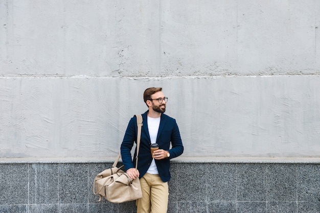 Retrato de hombre de negocios atractivo con anteojos sosteniendo un vaso de papel y una bolsa de transporte mientras está de pie cerca de la pared