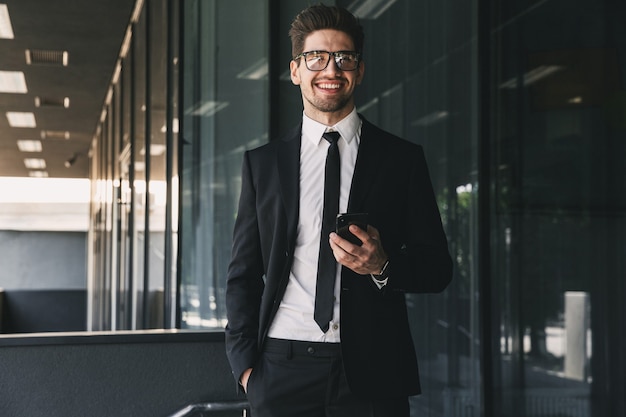 Foto retrato de hombre de negocios atractivo vestido con traje formal de pie fuera del edificio de cristal y sosteniendo un teléfono móvil