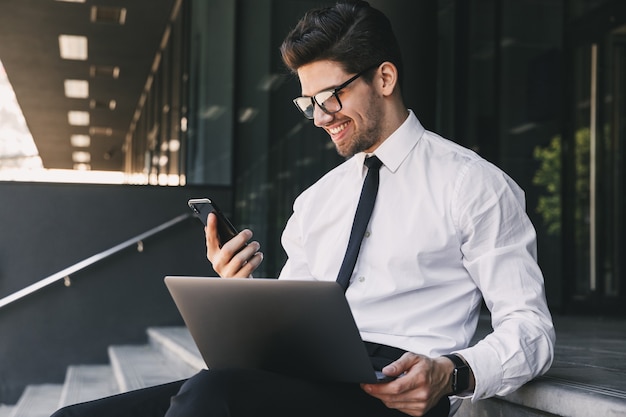 Retrato de hombre de negocios atractivo vestido con traje formal sentado fuera del edificio de cristal con ordenador portátil y sosteniendo smartphone