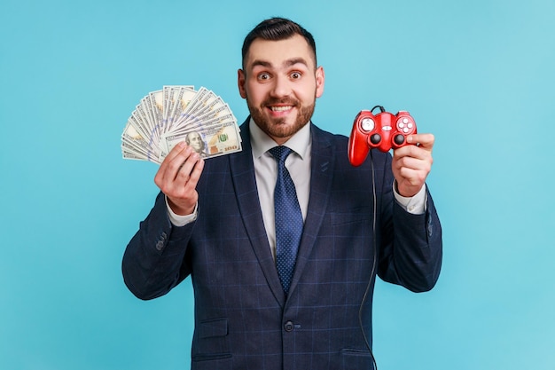 Retrato de un hombre de negocios barbudo sonriente con traje oscuro de estilo oficial sosteniendo en las manos billetes de dólar y joystick rojo mirando a la cámara Foto de estudio interior aislada en fondo azul