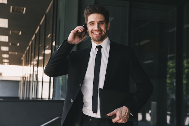 Foto retrato de hombre de negocios caucásico vestido con traje formal caminando fuera del edificio de cristal y hablando por teléfono móvil