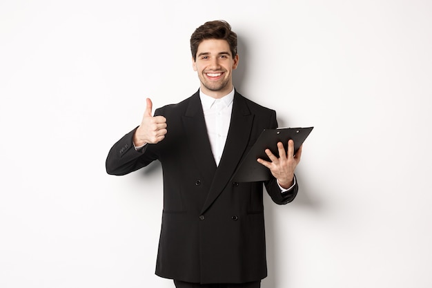 Retrato de hombre de negocios confiado en traje negro, sosteniendo el portapapeles con documentos y mostrando el pulgar hacia arriba en señal de aprobación, elogia el buen trabajo, de pie contra el fondo blanco.