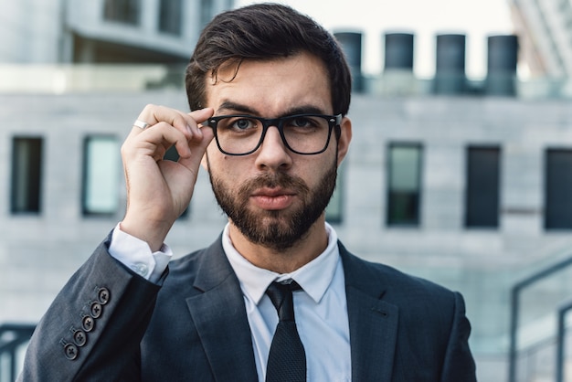 El retrato de un hombre de negocios está tocando las lentes en un traje clásico contra un edificio de oficinas.