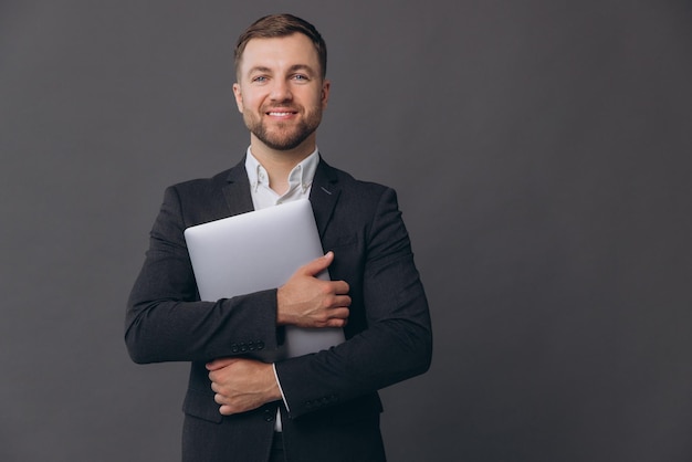 Retrato de un hombre de negocios exitoso y confiado en un traje gris trabajando con una computadora portátil y sonriendo feliz ante la cámara de pie sobre un fondo gris