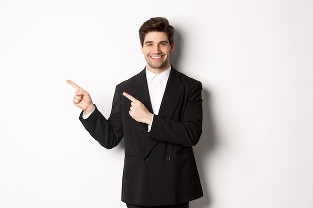 Retrato de hombre de negocios exitoso y guapo en traje, señalando con el dedo en la esquina superior izquierda y sonriendo, mostrando publicidad, de pie sobre fondo blanco.