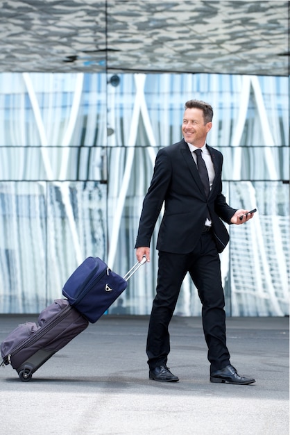 Retrato de hombre de negocios feliz caminando con equipaje y teléfono móvil