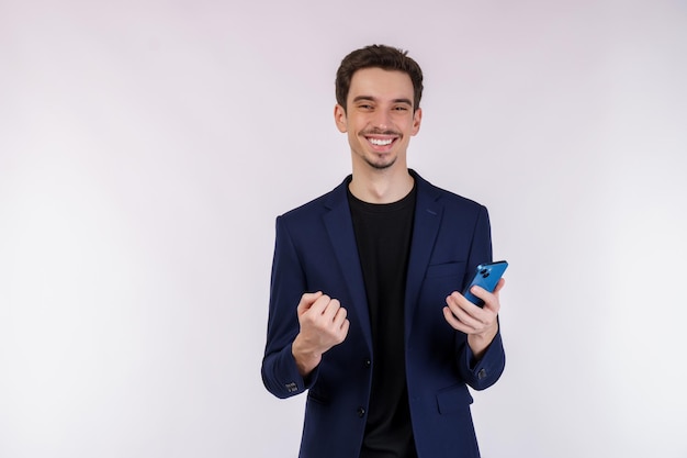 Foto retrato de un hombre de negocios feliz que usa un teléfono inteligente y hace un gesto de ganador apretando el puño sobre fondo blanco