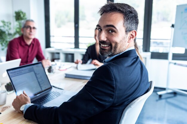 Retrato de hombre de negocios guapo está mirando a cámara mientras sus colegas están trabajando en lugar de coworking.