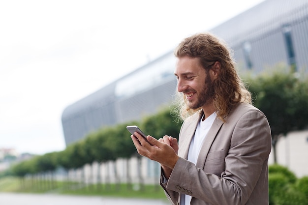 Retrato de hombre de negocios guapo rizado en ropa casual sosteniendo teléfono inteligente y sonriendo. Gerente exitoso usando aplicaciones de teléfono móvil, mensajes de texto, navegación por Internet, mirando el teléfono cerca del centro de negocios.