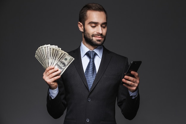 Retrato de un hombre de negocios guapo seguro con traje aislado, mostrando billetes de dinero, mediante teléfono móvil