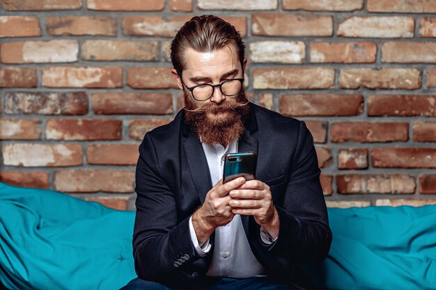 Retrato de hombre de negocios importante en gafas, con barba y bigote sosteniendo y enviando mensajes de texto en el teléfono