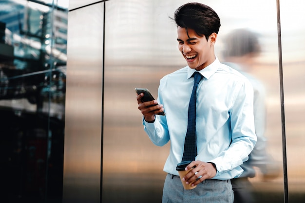 Retrato de un hombre de negocios joven feliz usando el teléfono móvil en la ciudad urbana. Estilo de vida de la gente moderna. De pie junto a la pared con una taza de café